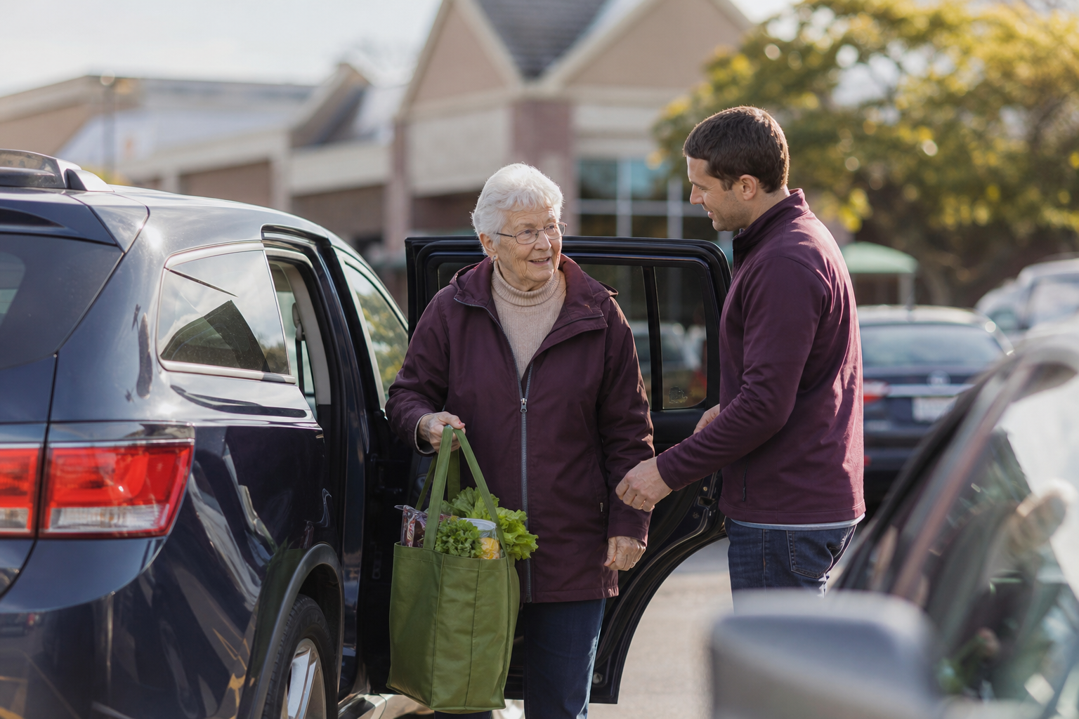 An elderly woman steps out of her adult son’s car at a grocery store entrance, smiling, holding a reusable bag, while he comes around to help.