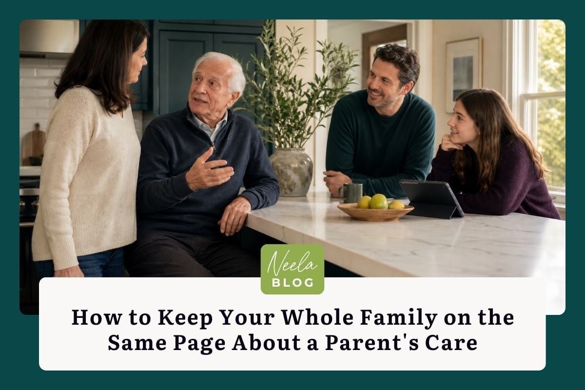 An older man in a navy sweater gestures as he speaks at a kitchen island, with his adult daughter, adult son, and teenage granddaughter gathered around him, listening and smiling.